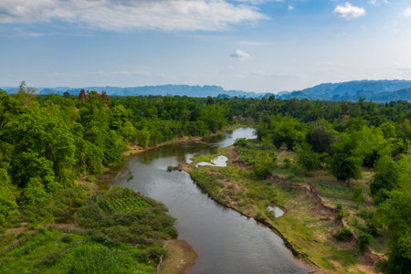A gentle river meanders through dense green vegetation and small cultivated plots in a countryside setting. Distant limestone mountains rise along the horizon beneath a soft blue sky, evoking a tranquil and natural atmosphere.の写真素材