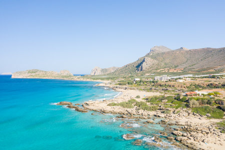 Bright turquoise water laps against a rugged, rocky coastline at Falasarna Beach, Crete. Sunlit hills and scattered buildings create a vibrant, inviting Mediterranean landscape.の写真素材