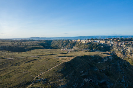 Wide aerial landscape showing the outskirts of Matera, Italy, with rugged grassy hills, rocky...