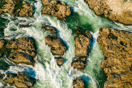 Aerial close-up reveals vibrant turquoise water surging between rugged golden rocks at Chutes de Khone in southern Laos. The dynamic patterns of the rapids highlight the textured stone and energetic river flow under clear daylight.の写真素材