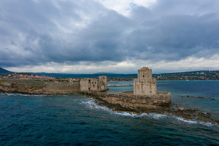 Historic Methoni Castle stands on a rugged stone outcrop surrounded by deep blue sea, with dramatic clouds overhead and the village visible in the distance.の写真素材