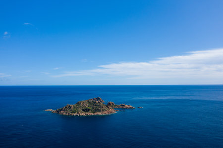 A solitary rocky island covered with green vegetation rises from the deep blue Mediterranean Sea beneath a vast, clear sky near Lotzorai, Sardinia. Bright sunlight and gentle clouds create a serene and expansive seascape.の写真素材