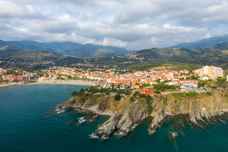 Aerial image of Banyuls-sur-Mer showing dramatic rocky cliffs meeting the turquoise Mediterranean Sea, with a vibrant village nestled along the shoreline and green mountains rising in the distance under a cloudy sky.の写真素材