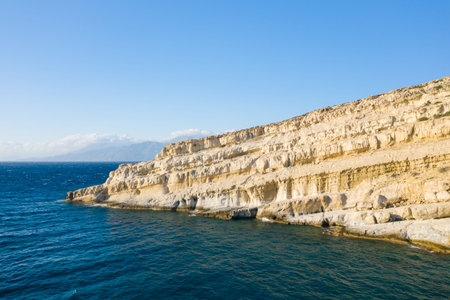 Sheer, pale limestone cliffs rise sharply from the vivid blue waters of the Mediterranean at Matala, Crete, under a clear sky. The textured rock faces are illuminated by bright sunlight, creating a striking coastal scene.の写真素材