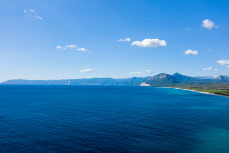 Wide aerial view of deep blue Mediterranean water meeting the long coastline and rugged mountains of Orosei under a bright sky with scattered clouds in Sardinia.の写真素材