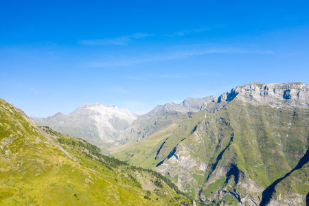 Expansive green valley flanked by rugged mountain slopes and distant sunlit summits under a clear blue sky near Gavarnie Gedre in the Pyrenees. Bright daylight reveals crisp textures and dramatic elevation changes.の写真素材