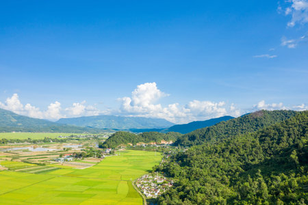Expansive view of a rural village nestled between lush green rice fields and densely forested hills,...