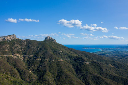 A winding road traverses rugged green mountains overlooking a sweeping bay and turquoise sea under a clear blue sky with scattered clouds in Sardinia, Italy. The scene combines dramatic elevation with coastal serenity and expansive views.の写真素材
