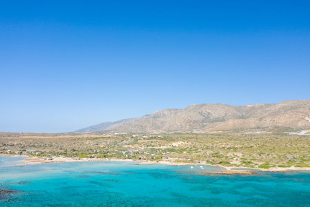 Bright turquoise water meets the rugged coastline of Elafonisi, Crete, with arid hills and a clear blue sky creating a vibrant, sunlit landscape. The open view highlights the natural textures and Mediterranean scenery.の写真素材