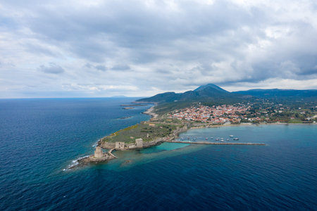 Aerial view of Methoni Castle extending into the sea, with a traditional Greek village and harbor nestled along the coastline under a dramatic cloudy sky.の写真素材