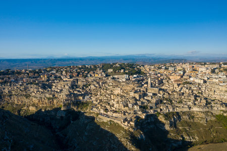 Aerial perspective of Matera, Italy, showing historic stone buildings clustered above a dramatic canyon, bathed in warm sunlight under a clear blue sky.の写真素材