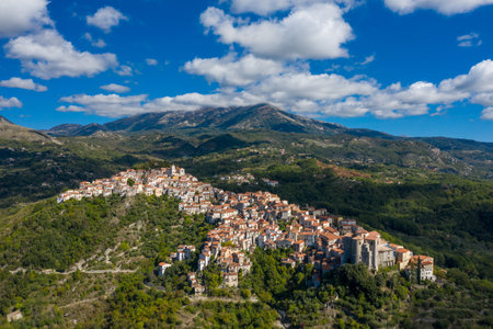 Aerial view of Rivello, Italy, featuring a dense cluster of terracotta-roofed houses nestled on a green hillside, framed by expansive mountains and a vivid blue sky with scattered clouds.の写真素材