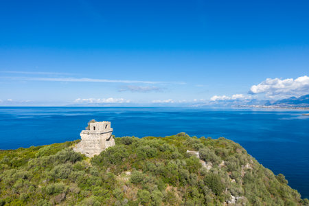 Ancient stone tower stands atop green vegetation on Isola di Cirella, surrounded by vivid blue Mediterranean waters under a clear sky. Bright daylight enhances the textures of the ruins and the expansive seascape.の写真素材