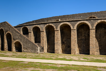 A row of ancient stone arches and a sloped stairway form the outer wall of the Roman amphitheatre in Pompeii, Italy. Bright sunlight reveals the volcanic masonry and geometric lines beneath a vivid blue sky, highlighting the historic architecture.の写真素材