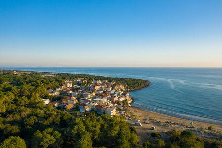 Aerial view of Arkoudi village with colorful houses nestled between green forest and sandy beach on the Ionian coast of Greece. Calm blue sea and clear sky create a peaceful Mediterranean atmosphere in this tranquil coastal settlement.の写真素材
