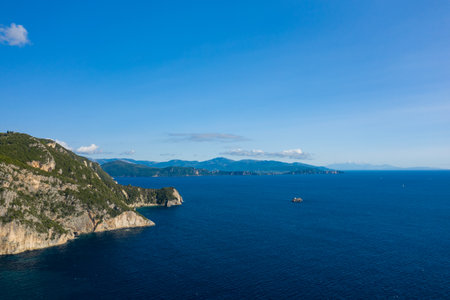 Wide view of the deep blue Ionian Sea meeting steep, rocky cliffs and green hills along the Epirus coastline near Parga, Greece. The clear sky and distant mountains create a tranquil Mediterranean seascape.の写真素材