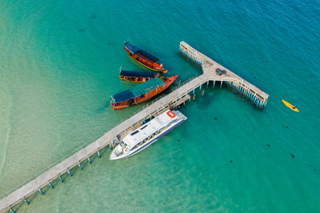 Aerial perspective shows several boats docked at a concrete pier extending into clear turquoise sea at Coconut Beach, Koh Rong, Cambodia. The scene features vibrant water, a yellow kayak, and subtle shadows beneath the surface, evoking a tranquil tropical setting.の写真素材