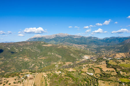 Drone view of rugged green hills and cultivated fields near Orgosolo, Sardinia, with a prominent mountain range under a vivid blue sky dotted with scattered clouds.の写真素材