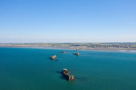 Aerial panorama of scattered concrete harbor remains stretching into calm turquoise sea near Asnelles, Normandy, with a distant sandy shoreline and village under a clear sky.の写真素材