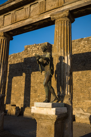 A bronze statue stands on a marble pedestal framed by weathered fluted columns at the Temple of Apollo in Pompeii, Italy. Warm sunlight casts dramatic shadows across the textured stonework, highlighting the classical forms of Roman architecture.の写真素材