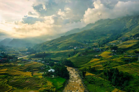 A meandering river flows between vibrant terraced rice fields and scattered rural houses in the green hills near Sapa, Vietnam. Dramatic clouds and golden sunlight create a serene, atmospheric landscape.の写真素材