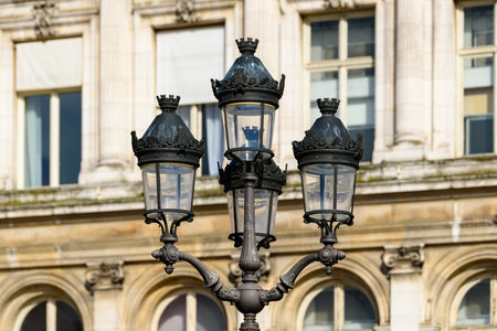 Elegant wrought iron street lamp featuring decorative crown details stands in front of the historic stone facade and arched windows of Hotel de Ville, illuminated by warm daylight.の写真素材