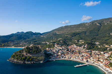 Aerial perspective of Parga in Epirus, Greece, showing a historic fortress on a rocky promontory above a colorful coastal village and turquoise bay. The scene is framed by densely forested hills and rugged mountains under a clear blue sky.の写真素材