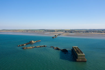 Aerial image of fragmented concrete blocks from a historic harbor stretching into calm turquoise sea near Asnelles, with sandy tidal flats and a distant village along the Normandy coastline under a clear blue sky.の写真素材