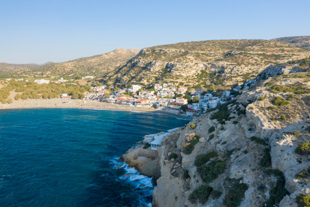Aerial view of the coastal village of Matala, Crete, nestled between golden limestone cliffs and the deep blue Mediterranean Sea. Sunlit hills and textured rock formations frame the tranquil bay and sandy beach.の写真素材