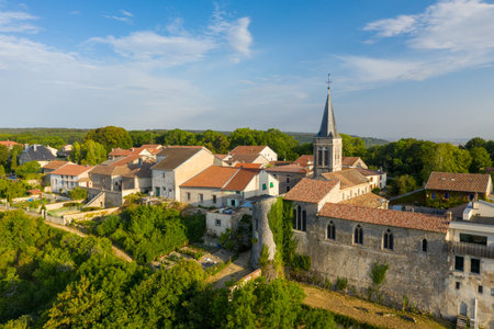 Elevated view of Hattonchatel village featuring a prominent stone church with a tall spire, traditional houses with red-tiled roofs, and lush greenery along the hillside. The late afternoon sunlight highlights rustic textures and the tranquil rural setting.の写真素材
