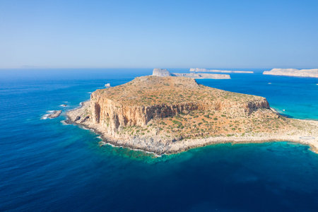 A rugged, sunlit peninsula juts into vivid blue Mediterranean waters at Balos Beach, Crete. The landscape features steep cliffs, sparse vegetation, and a tranquil, expansive seascape under a clear sky.の写真素材
