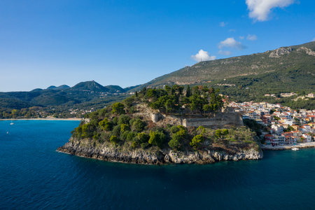 Aerial view of the historic castle ruins surrounded by pine trees atop a rocky hill above the turquoise waters and colorful houses of Parga, Greece. The Ionian coastline, forested hills, and distant mountains create a scenic Mediterranean landscape under a clear blue sky.の写真素材