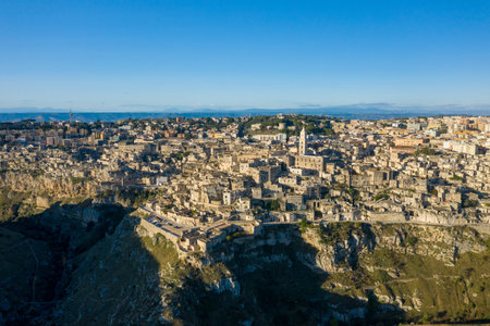 Wide aerial perspective of Matera, Italy, reveals ancient stone buildings cascading across dramatic cliffs under a clear blue sky, with sunlight highlighting the rugged terrain and layered architecture.の写真素材
