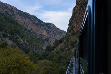 A narrow-gauge train travels along a bridge through the dramatic limestone cliffs and dense greenery of Vouraikos Gorge near Kalavrita, Greece. The scene features rugged mountain slopes, forested hillsides, and a tranquil sky.の写真素材