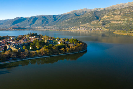 Aerial perspective of the historic peninsula of Ioannina surrounded by calm waters of Lake Ioannina, with the Pindus mountains forming a dramatic backdrop. The image features medieval walls, lush greenery, and red-tiled rooftops under clear daylight in Epirus, Greece.の写真素材