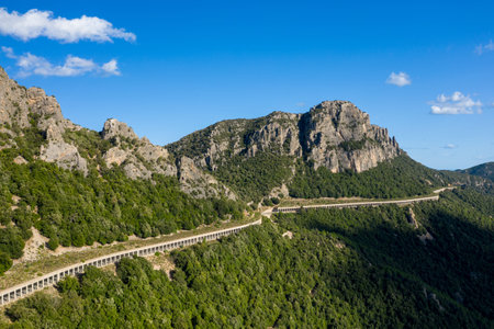 Elevated viaduct snakes along a lush green mountainside near Baunei and Dorgali in Sardinia, with dramatic rocky peaks rising under a clear blue sky.の写真素材