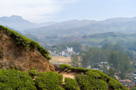 A rugged rocky outcrop with lush green tea bushes overlooks the town of Munnar, India, with rolling...