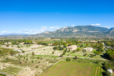Wide aerial panorama of the ancient archaeological site of Paestum, Italy, featuring expansive stone ruins and two well-preserved Greek temples set amid green fields. The scene is framed by distant mountains and a vivid blue Mediterranean sky with scattered clouds.の写真素材