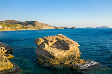 Large angular sandstone rock rises from clear turquoise sea at Triopetra beach, Crete, with rugged...