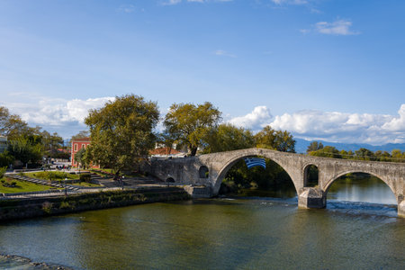 Wide view of the historic arched stone bridge in Arta, Greece, spanning a calm river. The scene includes a riverside park, mature trees, and a red building under a blue sky with scattered clouds.の写真素材