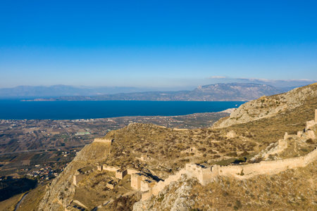 Ancient fortress remains stretch along the rugged slopes of Acrocorinth, overlooking patchwork farmland and the vivid blue Corinthian Gulf under a clear sky in Greece.の写真素材