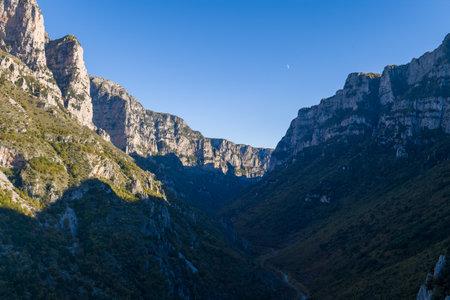 Wide view captures the towering limestone cliffs and deep shadowed valley of Vikos Gorge in Epirus, Greece. Crisp evening light highlights the dramatic rock faces and dense green slopes beneath a clear blue sky.の写真素材