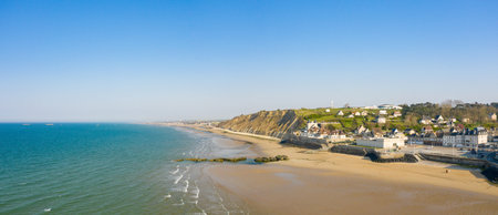 Bright coastal scene with sandy beach, turquoise sea, and a charming village set against steep grassy cliffs in Arromanches-les-Bains. Clear sky and gentle sunlight create a serene and inviting atmosphere.の写真素材
