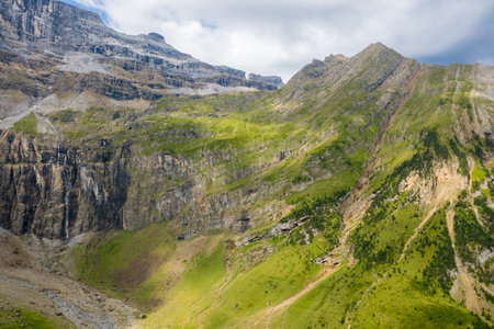 Expansive view of steep, rocky mountainsides with lush green slopes and dramatic cliff faces in Cirque de Gavarnie, illuminated by patches of sunlight under a partly cloudy sky.の写真素材