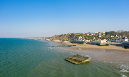 Expansive sandy shoreline meets turquoise sea at Arromanches-les-Bains, with dramatic cliffs and a historic concrete structure in the shallow water. Clear blue sky and soft sunlight create a bright, inviting coastal scene.の写真素材
