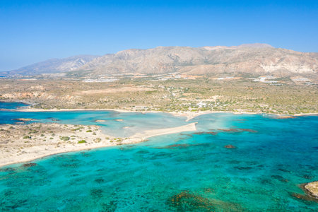 Aerial view of Elafonisi beach in Crete, Greece, with a striking sandbar stretching between turquoise shallows and rugged, sunlit hills. The tranquil scene features vivid blue water, soft sandy textures, and open Mediterranean landscape.の写真素材