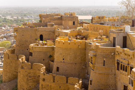 Golden sandstone bastions and terraces of Jaisalmer Fort rise above the city, featuring crenellated walls, sunlit textures, and scattered greenery against an arid landscape.の写真素材