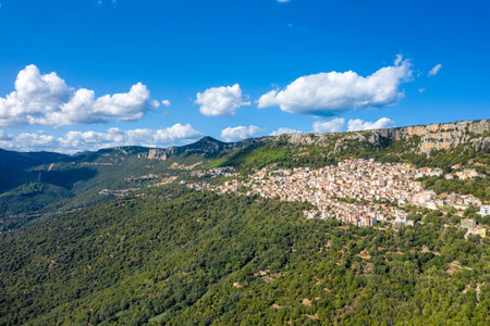 Aerial view of Baunei perched on a densely forested slope beneath towering limestone cliffs in Sardinia, Italy. Bright sunlight and scattered clouds create a vivid Mediterranean atmosphere.の写真素材