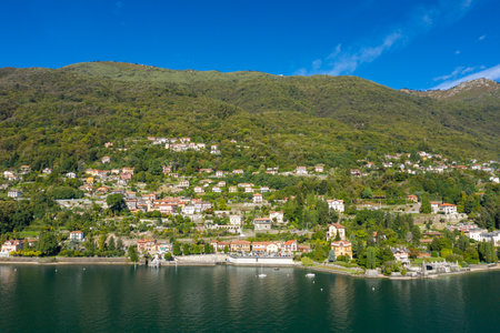 Panoramic view of a colorful hillside village overlooking the calm waters of Lake Maggiore in northern Italy. The scene features traditional houses nestled among lush green forest beneath a clear blue sky, creating a tranquil lakeside landscape.の写真素材