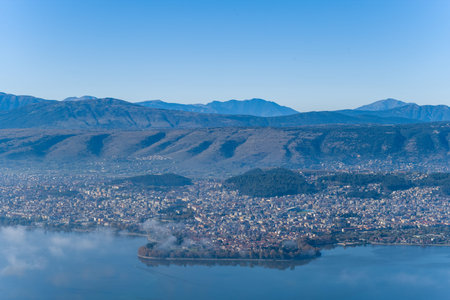 A wide panoramic view captures the city of Ioannina and its island on Lake Ioannina, set against the rugged slopes of the Pindus mountains under a clear blue sky in Epirus, Greece. Wisps of mist drift above the tranquil water, highlighting the natural textures and urban landscape.の写真素材